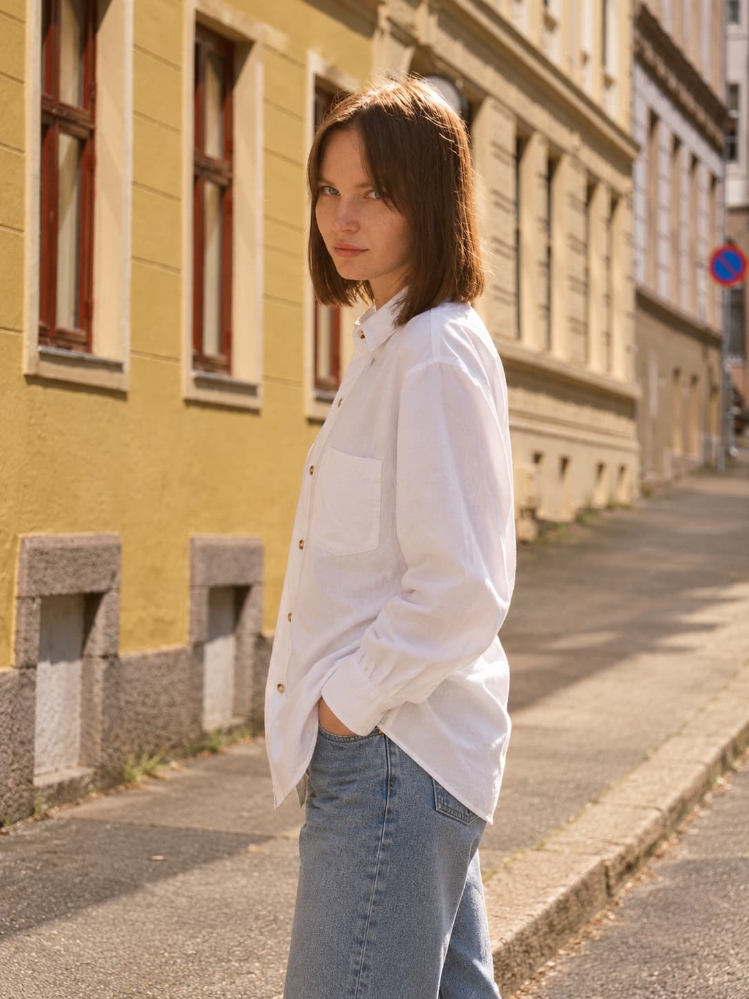 Nora in a white linen shirt with a chest pocket and light-wash jeans on a sunny street, looking over her shoulder